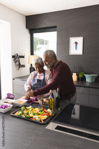 Happy senior biracial couple embracing and preparing vegetables in kitchen at home, copy space