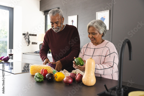 Happy senior biracial couple unpacking bags of vegetables in kitchen at home