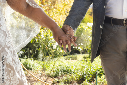 Midsection of senior biracial bride and groom holding hands at sunny outdoor wedding ceremony