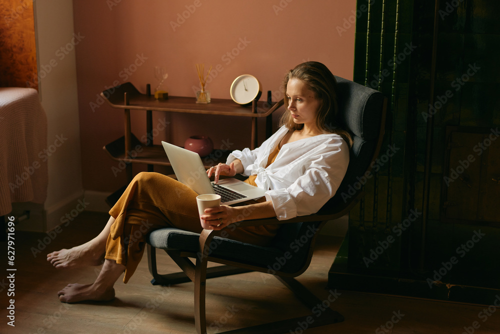 Relaxed woman sitting and watching something on her laptop with a cup of coffee in her hand Relaxed woman sitting and watching something on her laptop with a cup of coffee in her hand