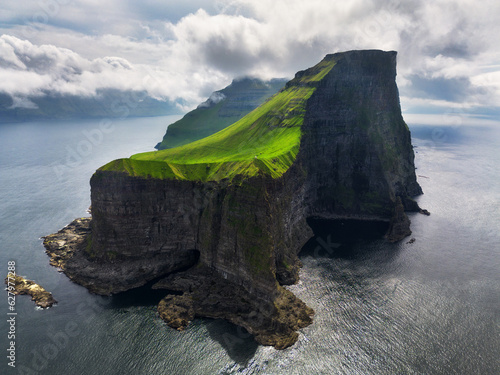  lighthouse sitting on top of a grassy hill close to a rocky black cliff looks over the tranquil sea on cloudy day. Breathtaking aerial view of a small green islet in Faroe Islands. photo