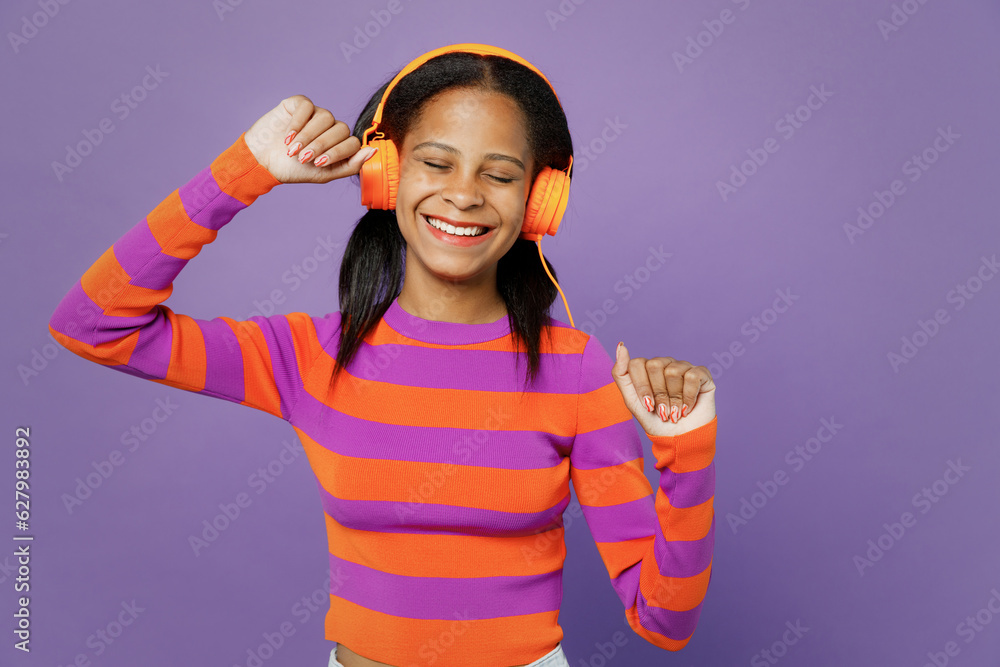 Little kid teen girl of African American ethnicity 15-16 years old wear striped orange sweatshirt listen to muisc in headphones dance isolated on plain purple background. Childhood lifestyle concept.