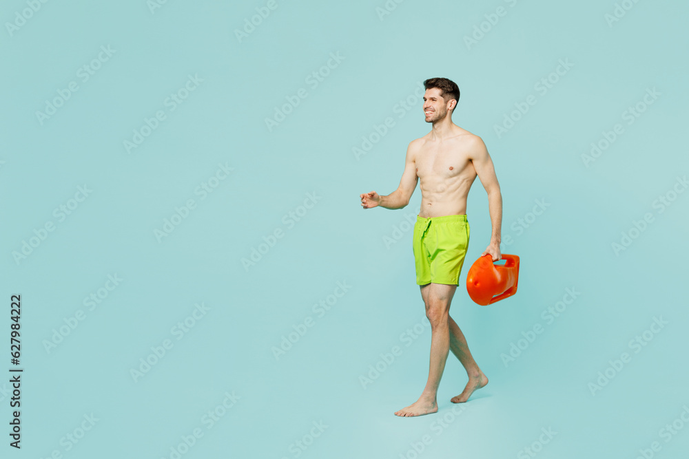 Full body side view young lifeguard man wear green shorts swimsuit relax near hotel pool hold lifebuoy walk go look aside isolated on plain blue background. Summer vacation sea rest sun tan concept.