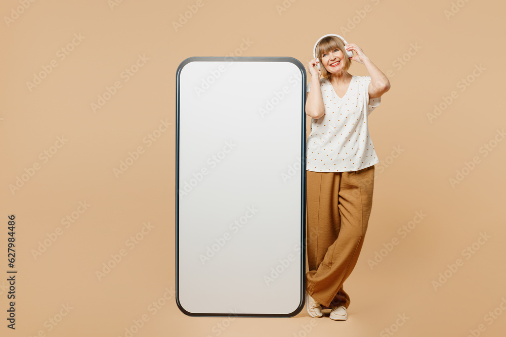 Full body blonde elderly woman 50s years old wear casual clothes big huge blank screen mobile cell phone smartphone with area listen to muisc in headphones isolated on plain light beige background. Full body blonde elderly woman 50s years old wear casual clothes big huge blank screen mobile cell phone smartphone with area listen to muisc in headphones isolated on plain light beige background.