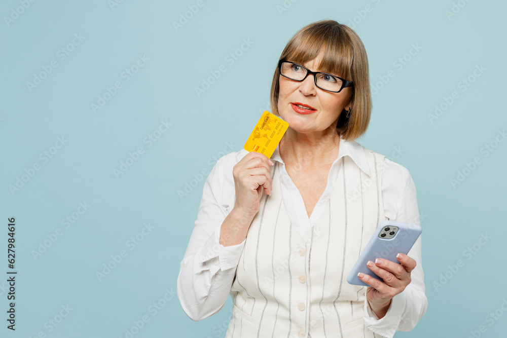 Employee business woman 50s wears white classic suit glasses formal clothes use mobile cell phone hold credit bank card shopping online isolated on plain blue background. Achievement career concept. Employee business woman 50s wears white classic suit glasses formal clothes use mobile cell phone hold credit bank card shopping online isolated on plain blue background. Achievement career concept.