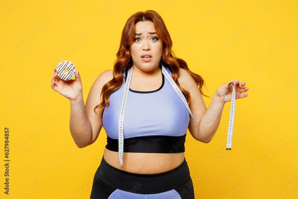 Young sad chubby overweight plus size big fat fit woman wear blue top warm up training hold in hand donut show measure tape isolated on plain yellow background studio home gym. Workout sport concept. Young sad chubby overweight plus size big fat fit woman wear blue top warm up training hold in hand donut show measure tape isolated on plain yellow background studio home gym. Workout sport concept.