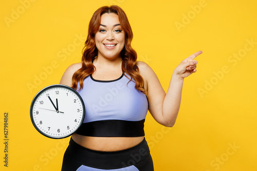 Young chubby overweight plus size big fat fit woman wears blue top warm up training hold in hands clock point finger aside isolated on plain yellow background studio home gym. Workout sport concept.