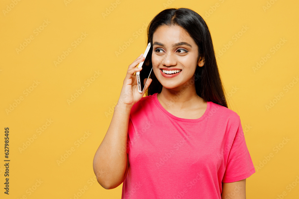 Young happy Indian woman wearing pink t-shirt casual clothes talking speak on mobile cell phone conducting pleasant conversation isolated on plain yellow background studio portrait. Lifestyle concept. Young happy Indian woman wearing pink t-shirt casual clothes talking speak on mobile cell phone conducting pleasant conversation isolated on plain yellow background studio portrait. Lifestyle concept.