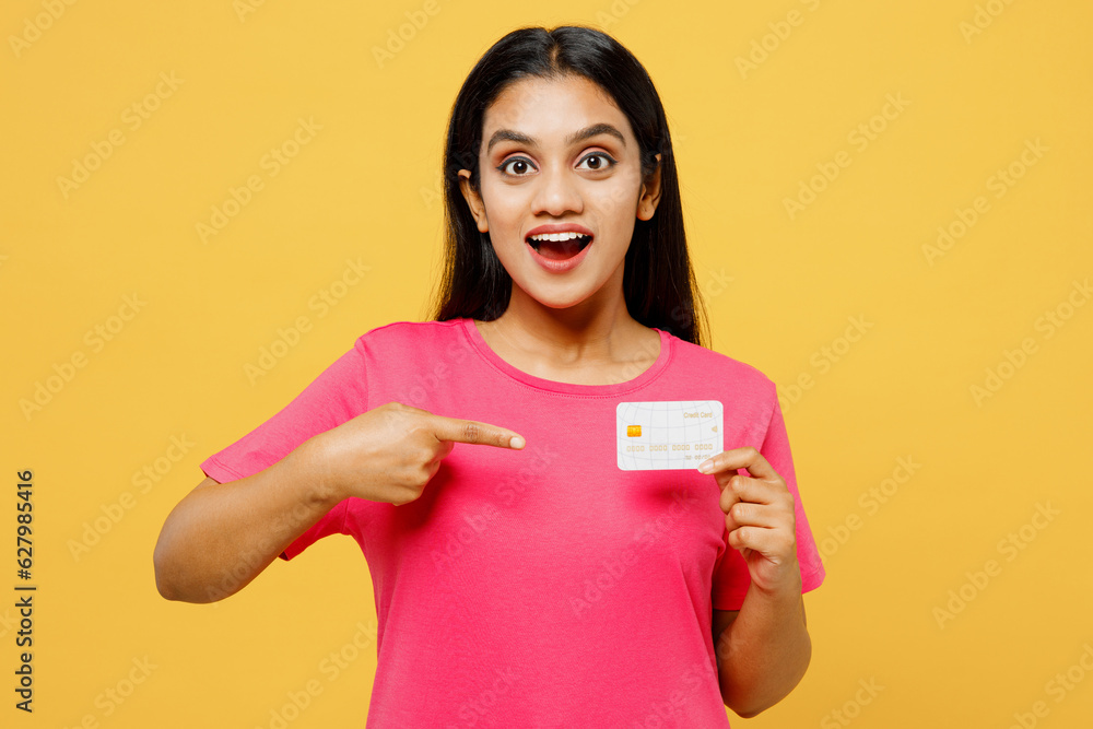 Young fun happy Indian woman wearing pink t-shirt casual clothes hold in hand point on mock up of credit bank card look camera isolated on plain yellow background studio portrait. Lifestyle concept. Young fun happy Indian woman wearing pink t-shirt casual clothes hold in hand point on mock up of credit bank card look camera isolated on plain yellow background studio portrait. Lifestyle concept.