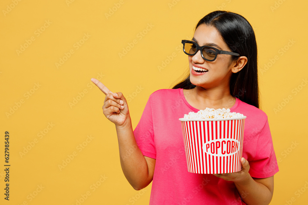 Young fun Indian woman in 3d glasses wearing pink t-shirt casual clothes watch movie film hold bucket of popcorn in cinema point index finger aside isolated on plain yellow background studio portrait.
