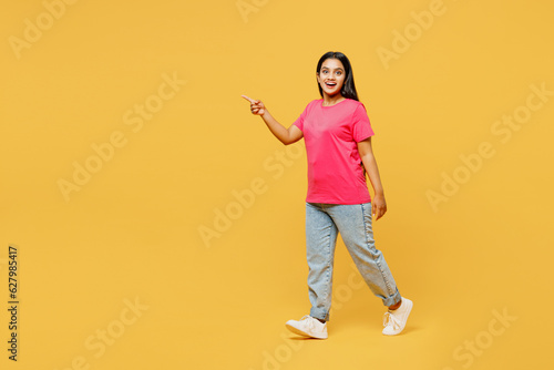 Full body side view young smiling cheerful happy Indian woman wear pink t-shirt casual clothes walking goign point finger aside isolated on plain yellow background studio portrait. Lifestyle concept.