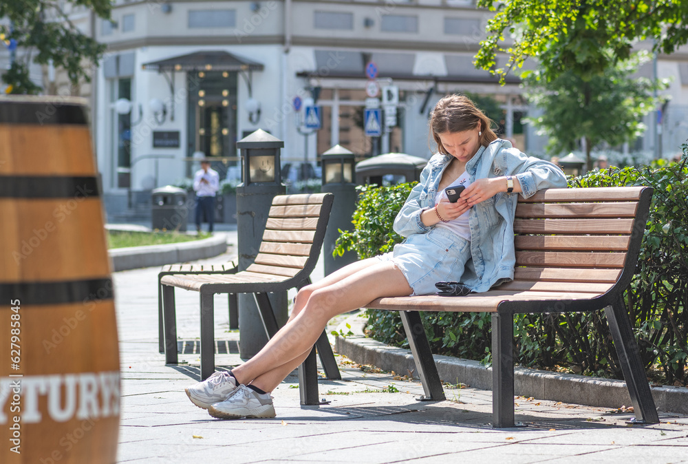 Beautiful Caucasian young girl sitting on the bench under the sun and checking the smartphone with a worried face 