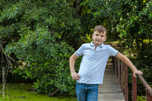 A boy standing on a bridge in the forest and looking at the camera with a smile. Rest in the village. Space for text.