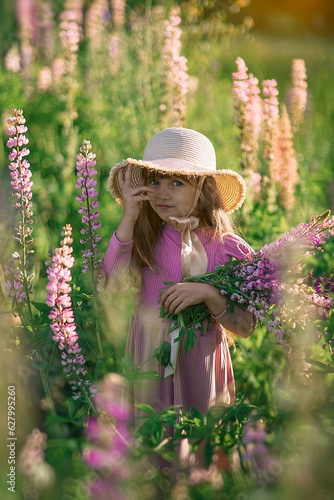 Beautiful little 7-old girl in a pink dress, straw hat with a bouquet of pink flowers in her hands at sunrise. Pretty child in summer in the bloomingfield holding a bunch of purple lupine at sundown