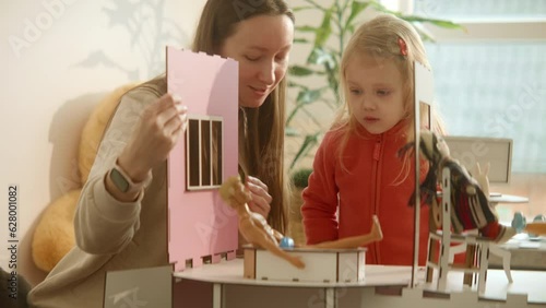 Mother and daughter in process of assembling wooden dolls house, playing with toys, looking for propper detail for building, family playtime delight, sunny weekend at bright cozy home. HQ 4k footage
