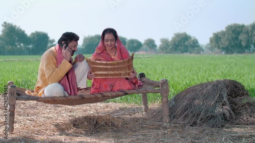 Indian village farmer talking on a mobile phone while his wife cleans food grain. Indian agricultural field in the background  village life. Happy Indian village family couple sitting in the shade ... photo