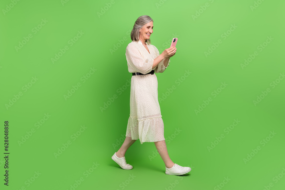 Full length photo of beautiful grandma walking hold telephone read news typing wear trendy white outfit isolated on green color background Full length photo of beautiful grandma walking hold telephone read news typing wear trendy white outfit isolated on green color background
