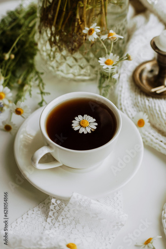Bright, light summer still life in a rustic style with a cup of coffee or tea, daisies and a lace ribbon on a white background. A chamomile flower floats in the drink