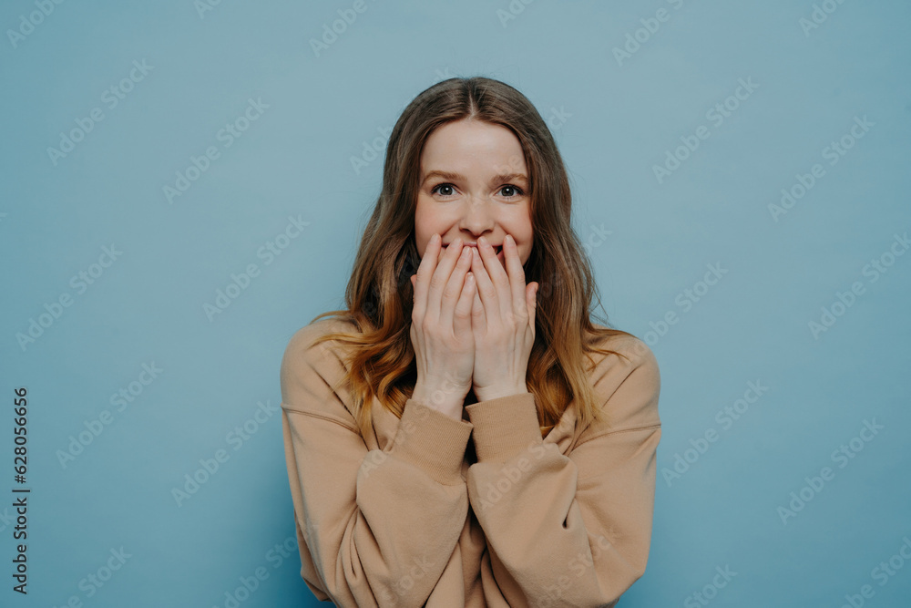 Amazed European teenage girl in casual clothes, laughing and covering mouth with hands, expressing positive emotions while posing against a blue wall in a studio. Amazed European teenage girl in casual clothes, laughing and covering mouth with hands, expressing positive emotions while posing against a blue wall in a studio.