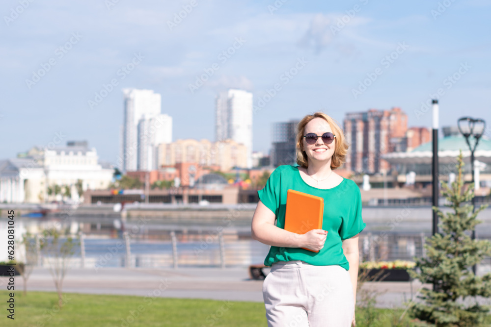 A smiling girl in sunglasses with a notebook or tablet in her hands on the city embankment. Break from work, study.