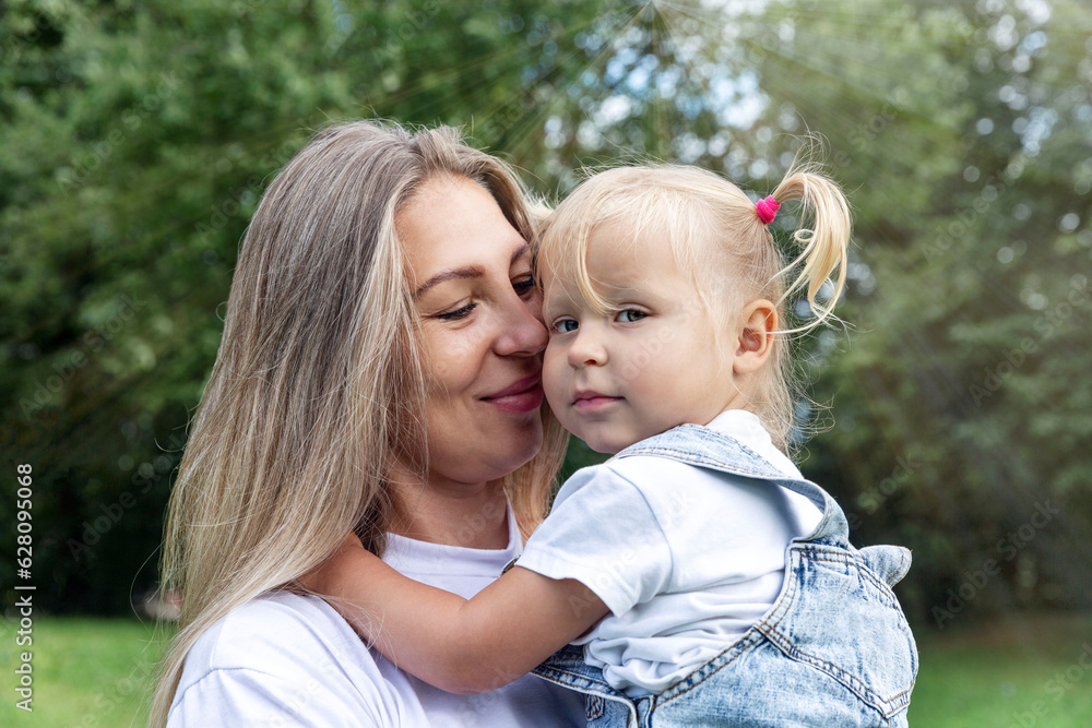 Laughing young woman with her little daughter in her arms in the park on a sunny summer day. Mom in a white T-shirt and jeans hugs her daughter in a denim sundress. Love and tenderness. Close-up. Laughing young woman with her little daughter in her arms in the park on a sunny summer day. Mom in a white T-shirt and jeans hugs her daughter in a denim sundress. Love and tenderness. Close-up.