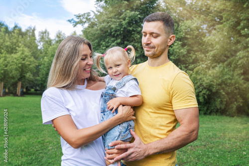 Happy parents with their little daughter in the park on a sunny summer day. Laughing young woman with a man and a child in jeans and t-shirts are hugging. Love and tenderness. © Анна Демидова
