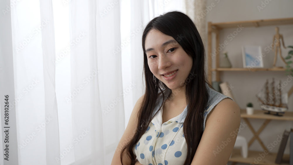 shoulder shot portrait of an attractive Asian woman look at the camera with smile near the window at home during the day. shoulder shot portrait of an attractive Asian woman look at the camera with smile near the window at home during the day.