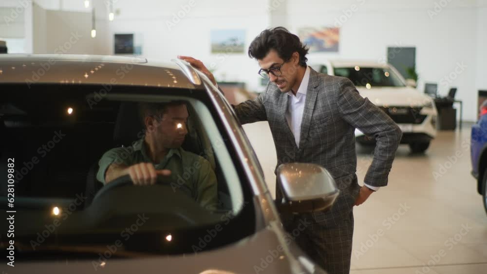 Portrait of serious male customer sitting on front seat of new car in dealership showroom touching steering wheel and having conversation with professional dealer. Concept of buying auto at showroom. Portrait of serious male customer sitting on front seat of new car in dealership showroom touching steering wheel and having conversation with professional dealer. Concept of buying auto at showroom.