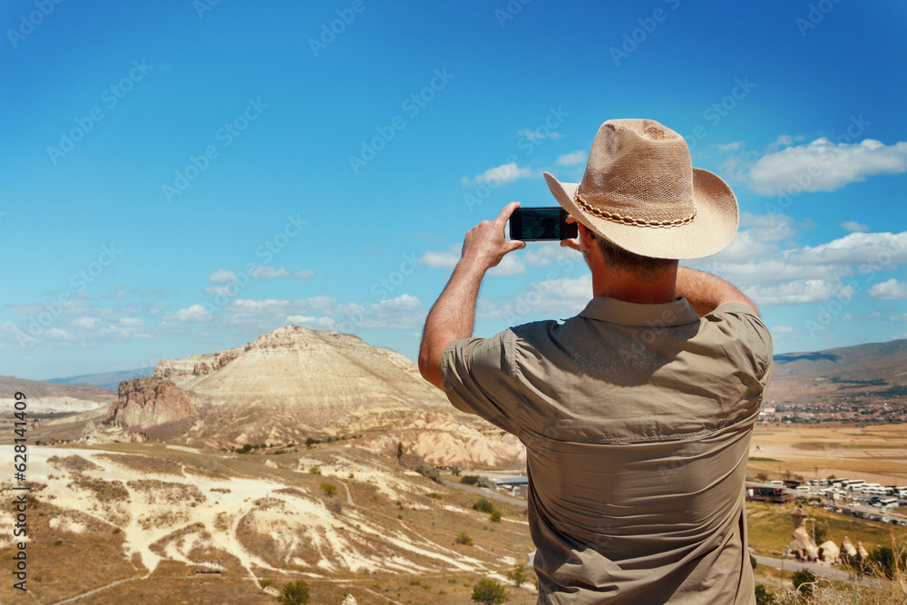 Man with shirt and cowboy hat taking photos with mobile phone during trekking in mountains