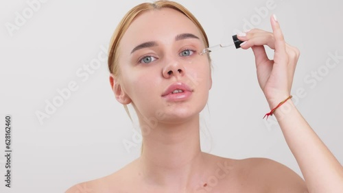 frame of young happy lady applying nourishing serum on face skin and rubbing it while smiling at camera on light background photo