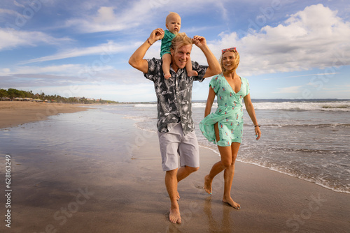 Happy Caucasian family of father, mother and son spending time on the beach. Baby sitting on daddy's neck. Summer vacation lifestyle concept. Travel to Asia. Seminyak beach, Bali