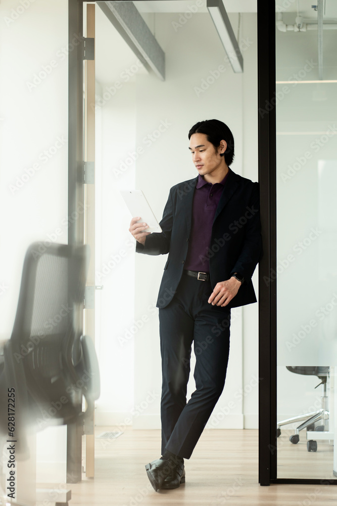 Handsome serious asian man holding digital tablet working online standing in modern office. Technology concept Handsome serious asian man holding digital tablet working online standing in modern office. Technology concept
