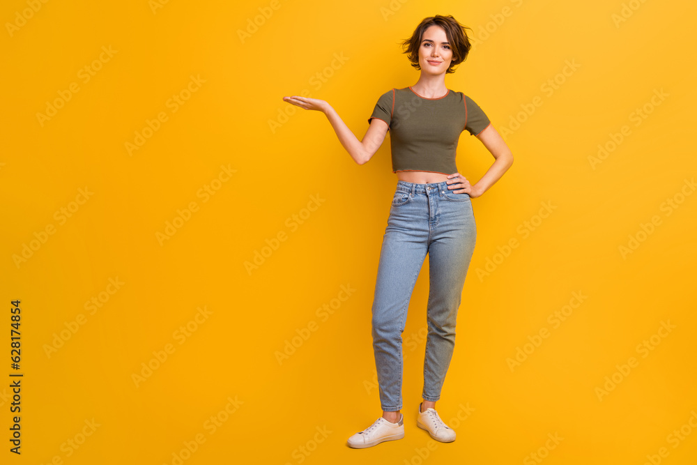 Full length photo of cute funky lady wear khaki t-shirt holding arm empty space isolated yellow color background Full length photo of cute funky lady wear khaki t-shirt holding arm empty space isolated yellow color background