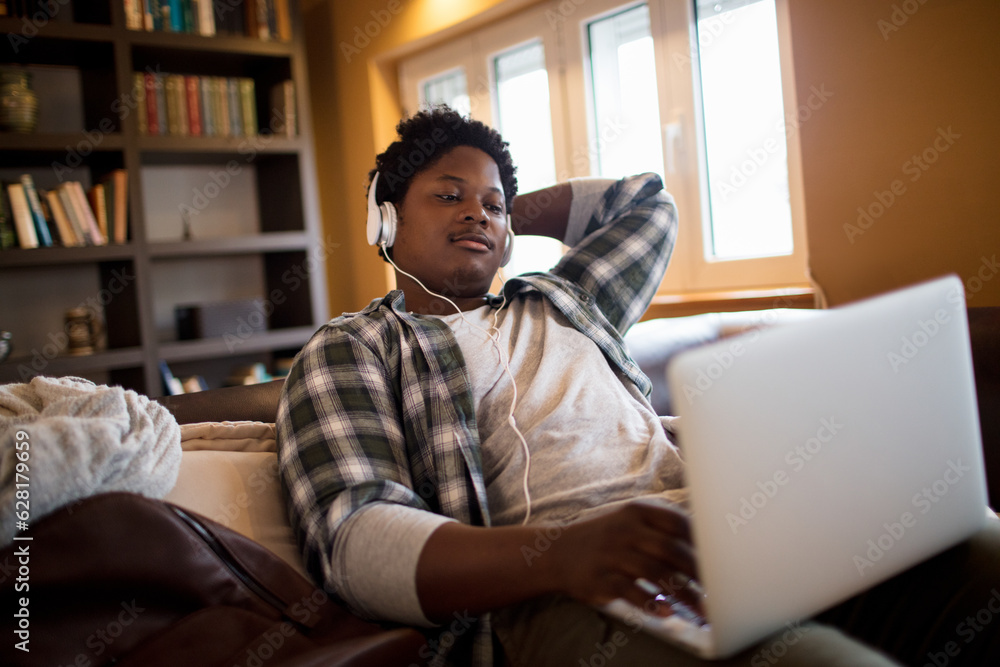 Young man of african ethnicity using a laptop at home while sitting on the couch and listening to music Young man of african ethnicity using a laptop at home while sitting on the couch and listening to music