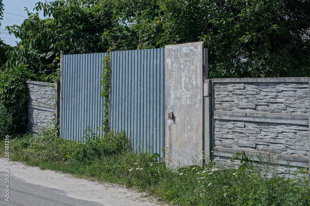 gray metal door and closed iron gate on a concrete fence wall overgrown with green vegetation on a rural street gray metal door and closed iron gate on a concrete fence wall overgrown with green vegetation on a rural street