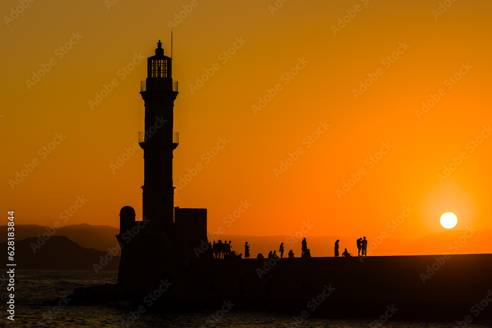 Silhouettes of people standing on a sea wall and lighthouse watching a beautiful orange sunset in summer