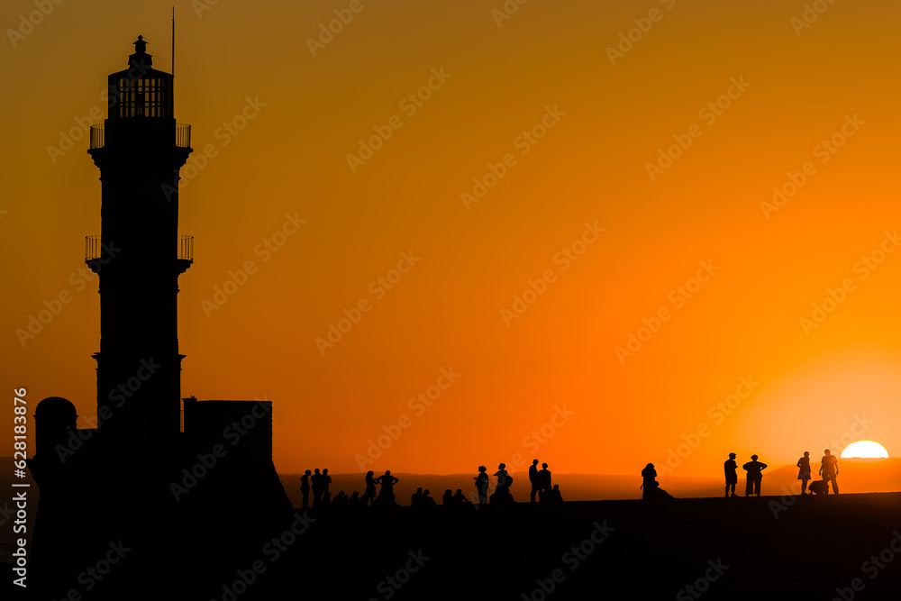 Groups of people standing on harbor wall next to a lighthouse watching a beautiful golden sunset (Chania, Crete) Groups of people standing on harbor wall next to a lighthouse watching a beautiful golden sunset (Chania, Crete)