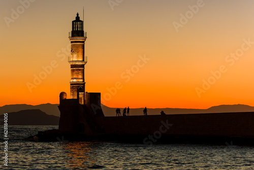 Groups of people standing on harbor wall next to a lighthouse watching a beautiful golden sunset (Chania, Crete)