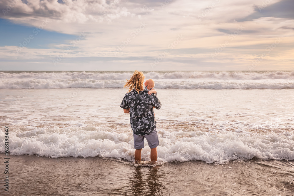 Caucasian father holding child in his arms. Happy childhood. Dad and baby son spending summer vacation on the beach. Horizon line. Ocean, waves, cloudy sky. View from back. Seminyak, Bali