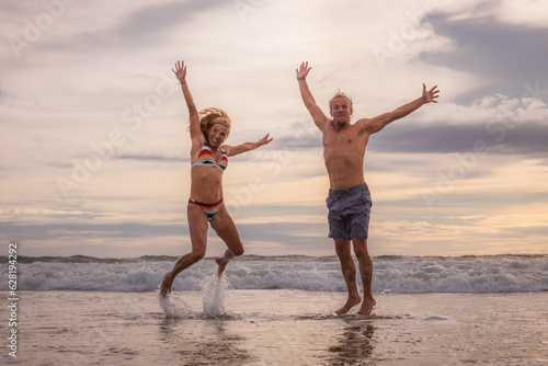 Happy Caucasian couple jumping on the beach. Motion body. Cheerful young man and woman enjoying life. Sunset time. Cloudy sky. Ocean with waves. Vacation in Asia. Bali, Seminyak