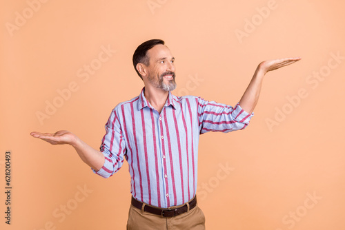 Photo of good mood man with gray white hair wear striped shirt palms show offer look empty space isolated on beige color background