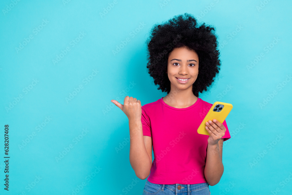 Photo of cheerful adorable small schoolgirl beaming smile hold smart p hone direct finger empty space promo isolated on teal color background