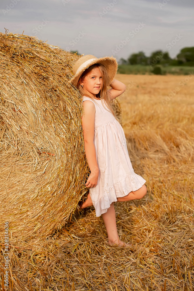 a beautiful blonde girl with long hair in a linen dress and a straw hat stands by a haystack in a mown field a beautiful blonde girl with long hair in a linen dress and a straw hat stands by a haystack in a mown field