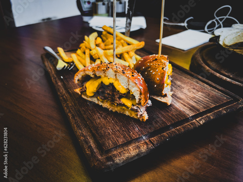 Cut in half huge triple burger with beef cutlets, fries onion rings, cherry, jalapeño and cheddar cheese on wooden cutting board over dark wooden background. Side view, close up