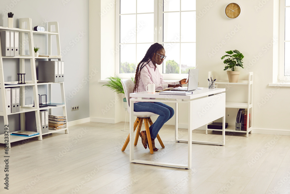 Side view portrait of a confident young business african american woman wearing casual clothes sitting at the desk on her workplace and working on a laptop at the modern white office.