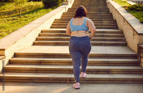 Back view of a overweight fat woman running up the stairs in the summer city park. Plus size girl wearing sportswear jogging outdoors. Weight loss, body positive, sport and fitness lifestyle concept.