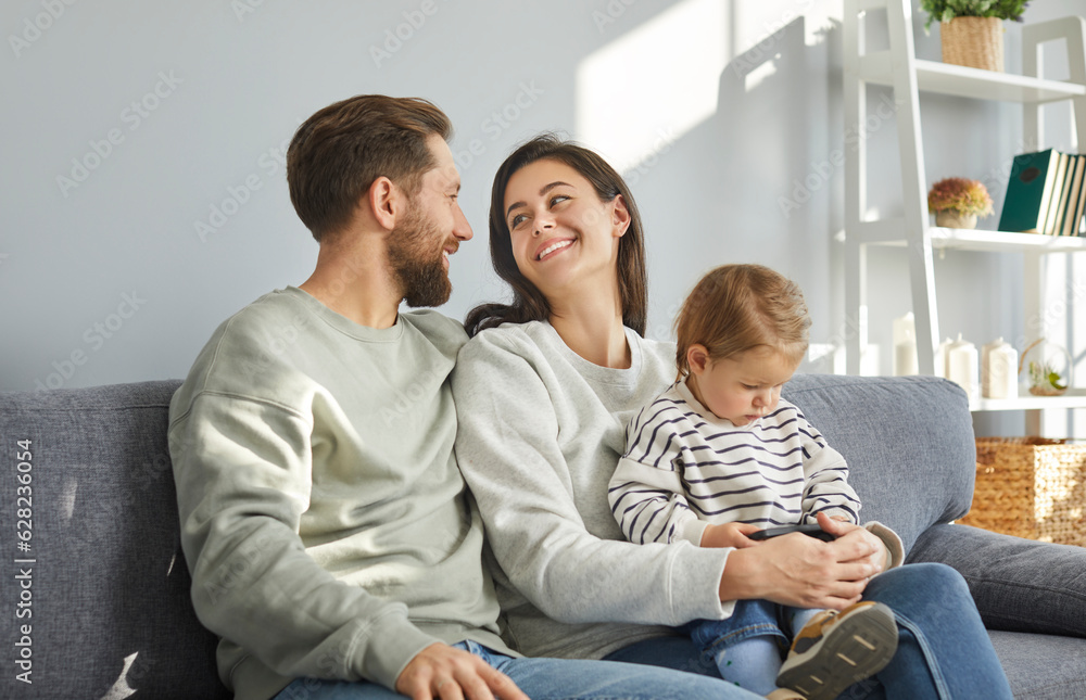 Happy parents and their toddler baby sitting on sofa together. Smiling parents looking at each other, kid sitting on knees of her mother looking at smartphone screen. Family spending time at home Happy parents and their toddler baby sitting on sofa together. Smiling parents looking at each other, kid sitting on knees of her mother looking at smartphone screen. Family spending time at home