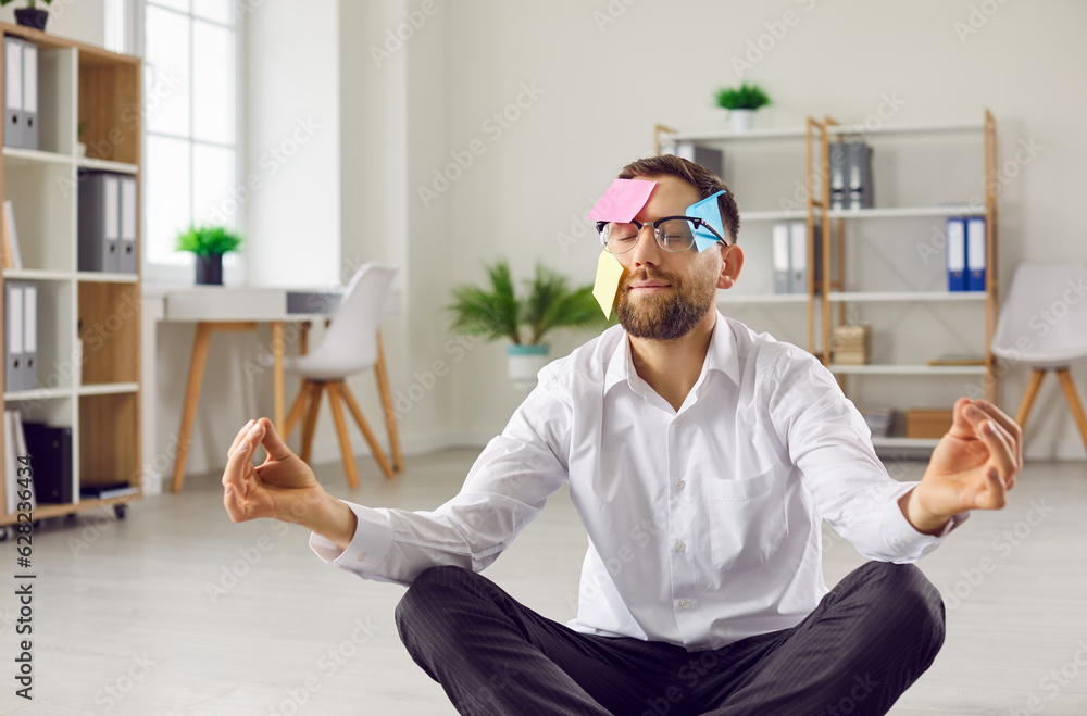 Funny business man or busy corporate employee with post it notes on face practising stress management, relaxing and meditating while sitting on floor in yoga pose, with office workplace in background - obrazy, fototapety, plakaty 