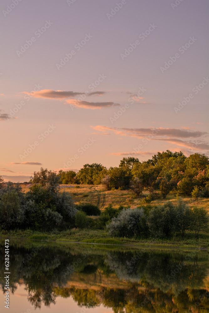 fields with hills on which green grass, bushes and trees grow, near a calm lake, against the backdrop of a bright sunset sky with clouds, during the golden hour fields with hills on which green grass, bushes and trees grow, near a calm lake, against the backdrop of a bright sunset sky with clouds, during the golden hour