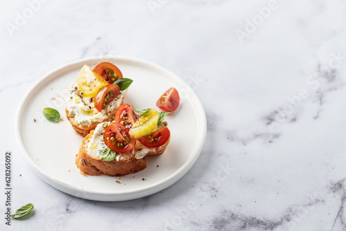Bruschetta sandwiches with tomatoes, cream cheese, olive oil and basil on a plate on white marble background, copy space. Traditional italian antipasti